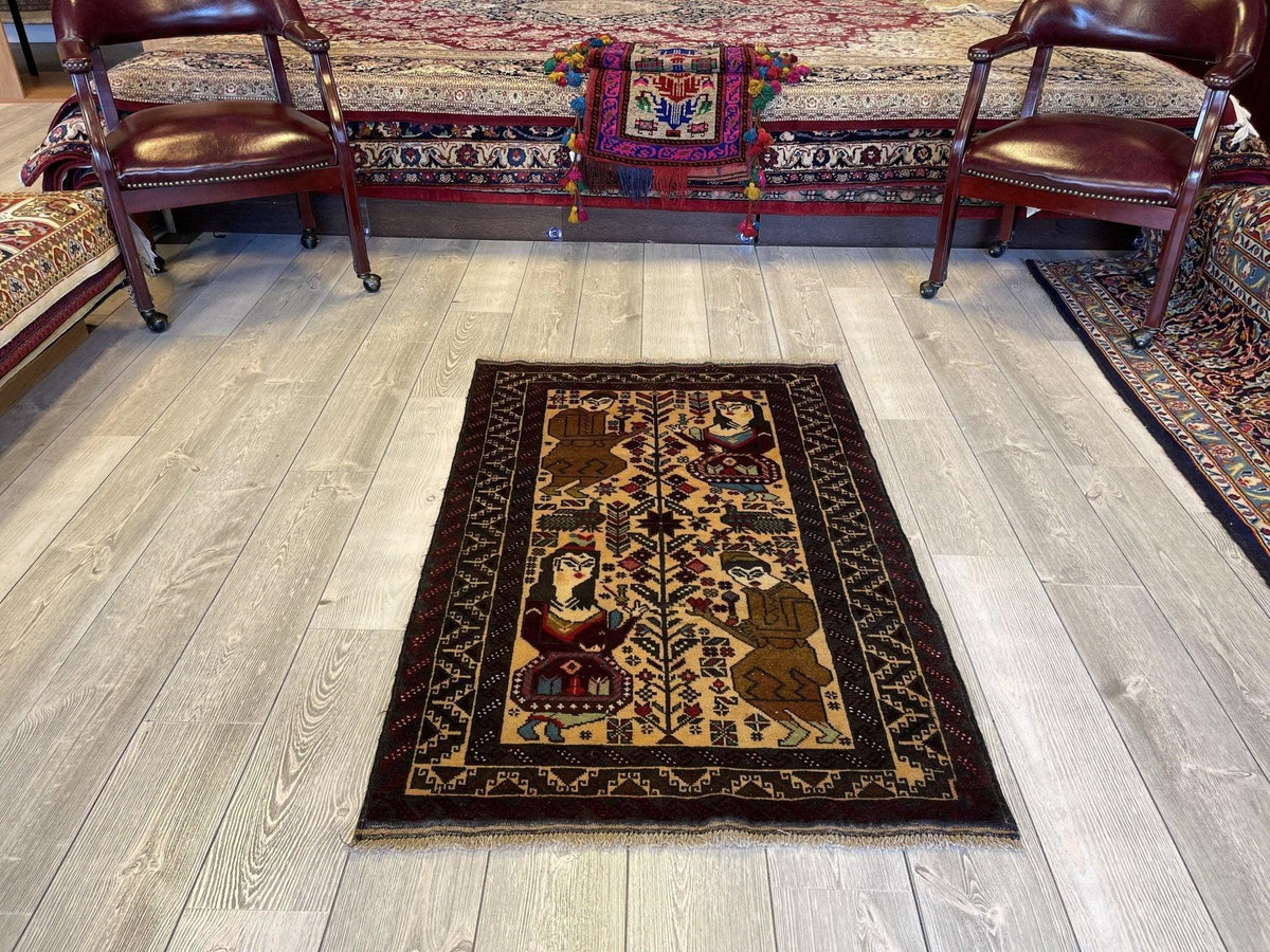 Small rectangular rug with a central tree motif flanked by stylized human and animal figures in brown, black, and beige tones on light wood flooring between two leather chairs.
