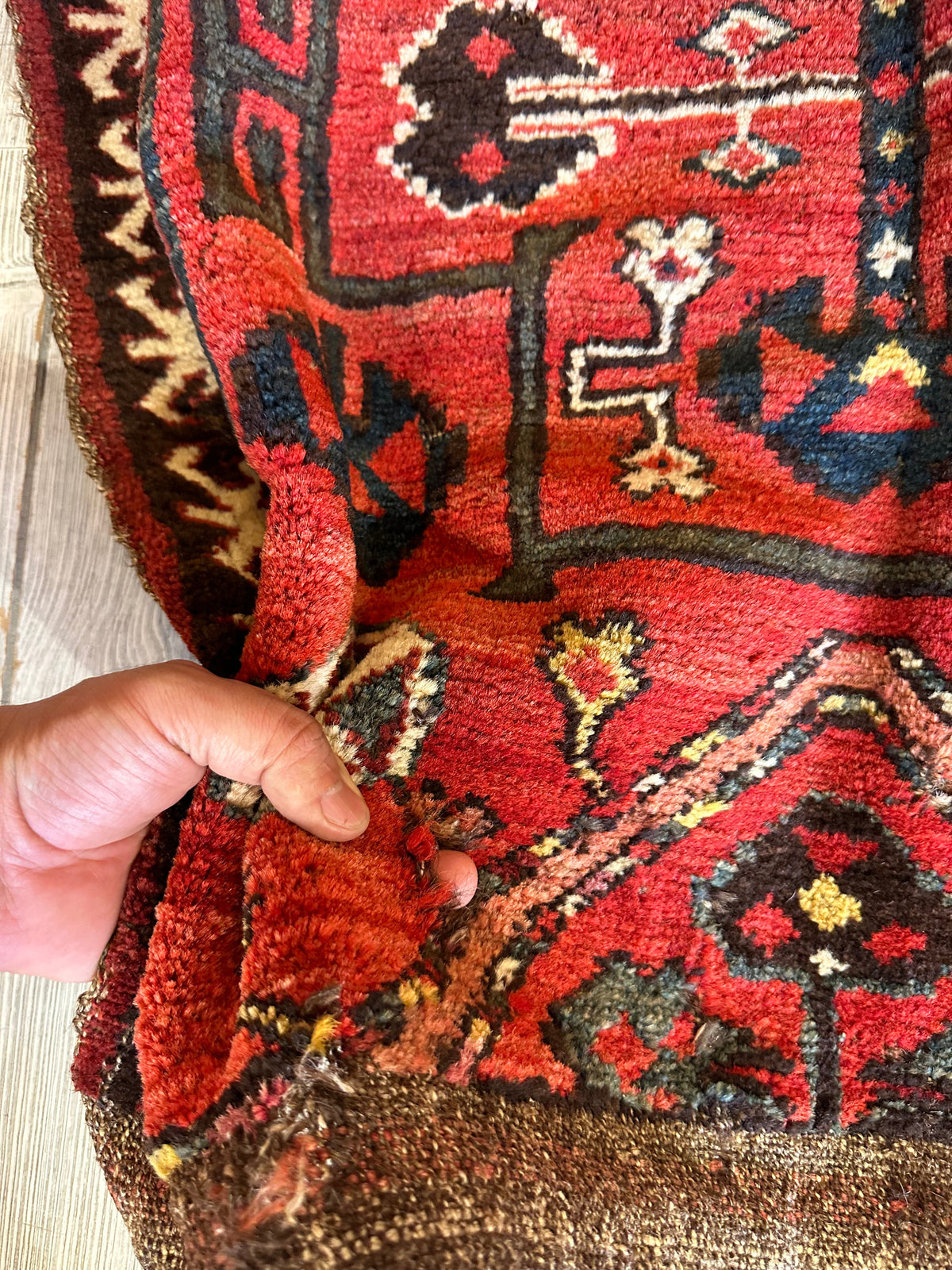 Hand holds antique tribal bagface textile featuring intricate geometric and floral motifs in red, black, blue, and beige tones over coarse woven backing.