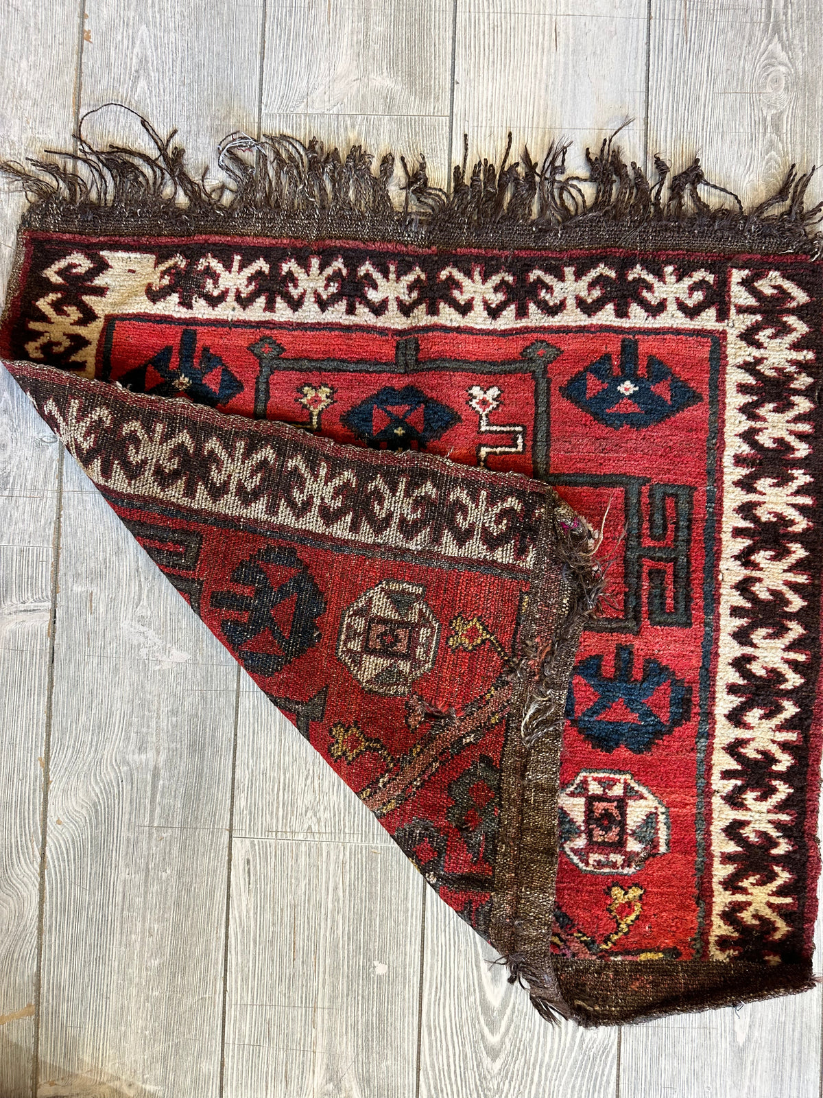 Antique tribal bagface with red, cream, and brown geometric patterns and dark blue accents, showing folded corner and fringed edge on light wood floor.