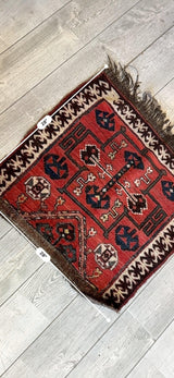 Square antique tribal bagface with red background, geometric patterns in dark blue, white, and brown, and fringed edge on wooden floor.