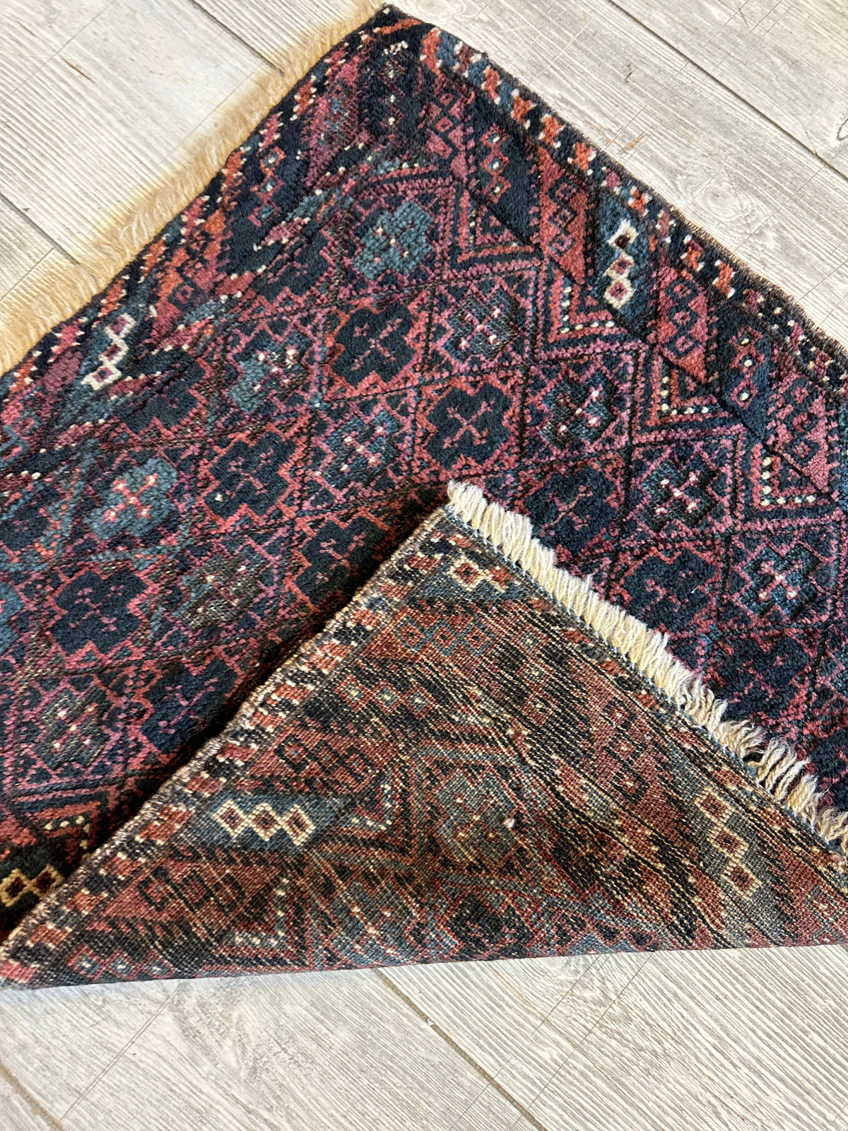 Antique Baluch tribal bag face with dark red and navy geometric patterns, showing a folded corner revealing a beige fringe and backside design.