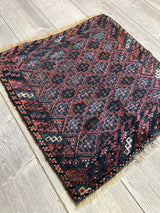 Square antique Baluch tribal bag face featuring dark blue and red geometric diamond patterns with intricate cross motifs and white accents on a wooden floor.