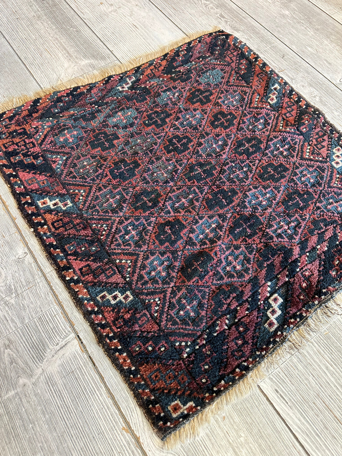 Square antique Baluch tribal bag face featuring dark blue and red geometric diamond patterns with intricate cross motifs and white accents on a wooden floor.