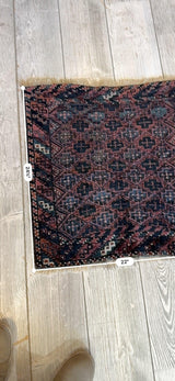 Rectangular antique tribal Baluch bag face with intricate dark red, black, and blue geometric patterns on a beige wooden floor, measuring 22 by 24 inches.