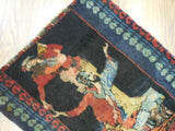 Small rectangular antique rug with central pictorial design featuring human and animal figures in red, blue, yellow, and green tones framed by patterned borders.