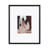 Black photo frame with beaded textured edges surrounding a centered photo of a small black and white dog wearing a red harness, standing among people wearing tan and pink shoes.