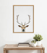 Framed canvas showing a frontal portrait of a deer with large antlers against a plain light background, hung above a wooden table with books and potted plants.