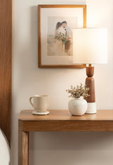 Wooden table with a ceramic mug, white vase holding small flowers, and a wooden table lamp with a white shade under a framed photo on a cream wall.