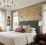 Bedroom with a green upholstered headboard against beige floral wallpaper featuring birds, cream bedding, and decorative pillows including a central mauve lumbar pillow.
