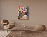 Bright impressionist painting of sunlit stone arches and stairs with pink and yellow buildings, green foliage, and blue windows on a canvas print hung above beige living room furniture.