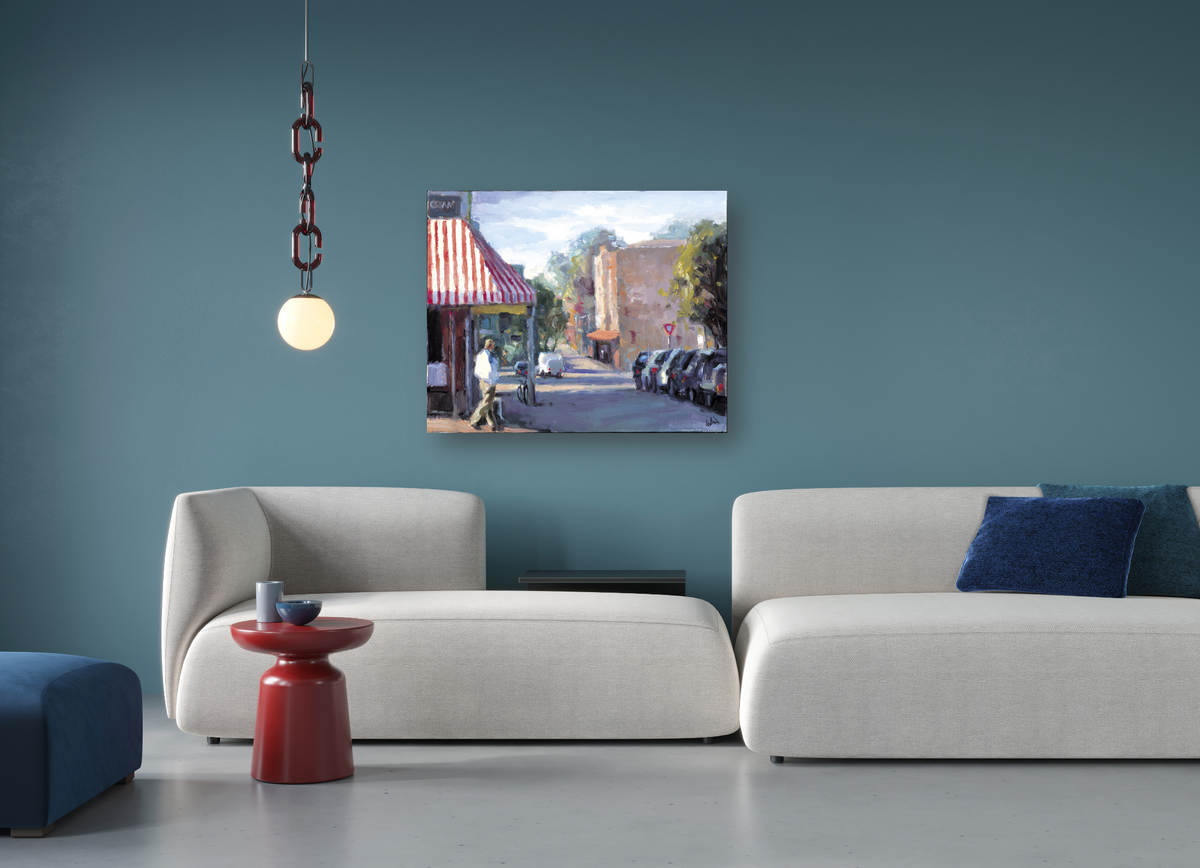 Artwork showing a sunlit urban street corner with a café, outdoor tables, and parked cars, displayed on a teal wall above modern cream sofas with blue accent pillows.