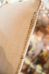 Close-up of a pillow with beige woven fabric on one side and a vintage rug fragment pattern in neutral tones on the edge, shown with a blurred warm background.