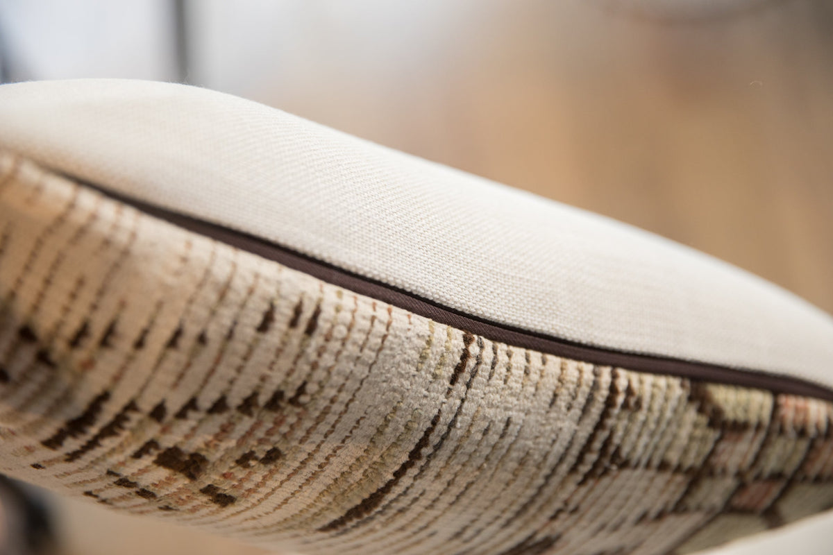 Close-up of a pillow with a textured beige fabric top and a vintage rug fragment pattern in muted earth tones on the side, separated by dark piping.