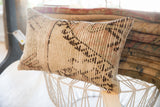 Rectangular pillow with a beige base and dark brown geometric patterns, resting on a white wire-frame stool with stacked folded rugs behind it.