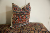Square pillow with intricate vintage-style rug pattern in red, blue, and cream colors, resting on a similarly patterned surface against a plain white background.