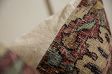 Close-up of a pillow showing beige fabric and a vintage rug fragment with red, brown, blue, and black pattern along one edge.