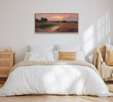 Framed landscape painting above bed showing soft pink and orange sunset over grassy foothills and a winding reflective stream.