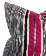 Close-up of a textured fabric pillow cover with horizontal stripes in berry pink, black, cream, and varying shades of gray, showing a corner and fold detail.