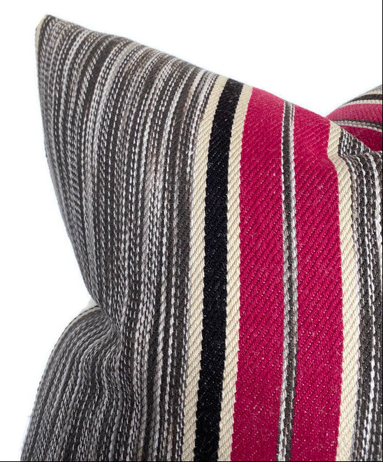 Close-up of a textured fabric pillow cover with horizontal stripes in berry pink, black, cream, and varying shades of gray, showing a corner and fold detail.