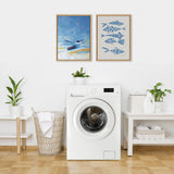 Two framed artworks above a white front-loading washing machine, one showing blue boats on a sandy shore and the other featuring stylized blue fish on beige background, in a minimalist laundry room.