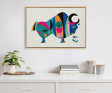 Framed canvas on a white wall featuring a geometric ox with bold colorful shapes in blue, orange, pink, black, and teal above a white cabinet with plants and books.