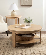 Round light wood coffee table with lower shelf holding a woven basket, potted plant and bowl on top in a neutral living room setting with matching side table and lamp.