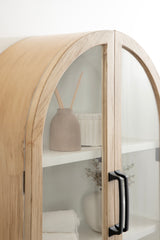 Curved wooden wall cabinet with glass arch doors and black handles, showing interior shelves with a beige vase, white dish, and folded towel.