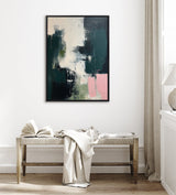 Abstract canvas painting with dark emerald green, blush pink, white, and black brushstrokes, framed in black and hung on a white wall above a bench with a light fabric throw and open book.