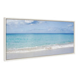 Framed horizontal canvas print showing a calm ocean with turquoise waves washing onto a sandy beach under a partly cloudy blue sky.