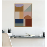Framed abstract canvas print with geometric shapes in muted earth tones, blue, and mauve hanging above a modern desk setup with books and accessories.