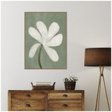 Framed canvas of a white floral illustration with a green stem on a muted green background hanging above a wooden dresser beside a tripod floor lamp and a wicker basket with flowers.