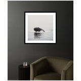Black and white framed photo of a humpback whale partially breaching in calm water, hanging on a dark gray wall above an olive green armchair and black side table.