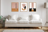 Three framed abstract art pieces above a white cushioned sofa, featuring geometric shapes in pink, black, and tan tones against a light gray wall with a floor lamp on the right and a potted plant on the left.