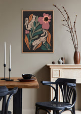 Framed canvas artwork showing two cream-colored leopards with black spots among large green leaves and a pink flower on a dark background in a cozy dining room.
