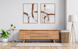 Two framed abstract canvas artworks with white backgrounds and curved thick brown brushstrokes, hanging above a low wooden sideboard in a minimalist living room setting.