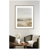 Framed abstract landscape print with soft beige and gray tones depicting fading fields under a cloudy sky, hanging above a dark armchair with a white throw and round wooden table with white vase.