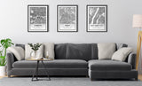 Three black and white framed city map prints of London, Paris, and New York City hanging above a dark gray sectional sofa with beige and light gray pillows in a minimalist living room.
