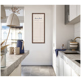 Tall, narrow framed fabric pinboard styled as a linen guest check form with blue text, hung on a white kitchen wall between two concrete countertops.