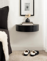 Black semi-circular floating wood side table mounted on white wall, holding a dark matte vase with light branches and two books, with black slippers on carpet below.