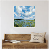 Square framed painting of a Texas bluebonnet field under a dynamic blue sky with swirling white clouds, hung above a wooden bench with a 'welcome home' pillow and open book.