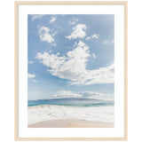 Framed photograph of a tranquil beach with gentle waves washing onto the sand, a distant island on the horizon beneath a bright blue sky with scattered white clouds.