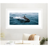 Framed photo of a dolphin leaping through deep blue ocean waves, backlit by soft natural light, displayed on a white wall above a beige chair with a brown throw.