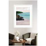Framed seascape art featuring turquoise water meeting a sandy beach with dark rocks and green cliff under a cloudy sky, hung above two gray armchairs and a wooden table.