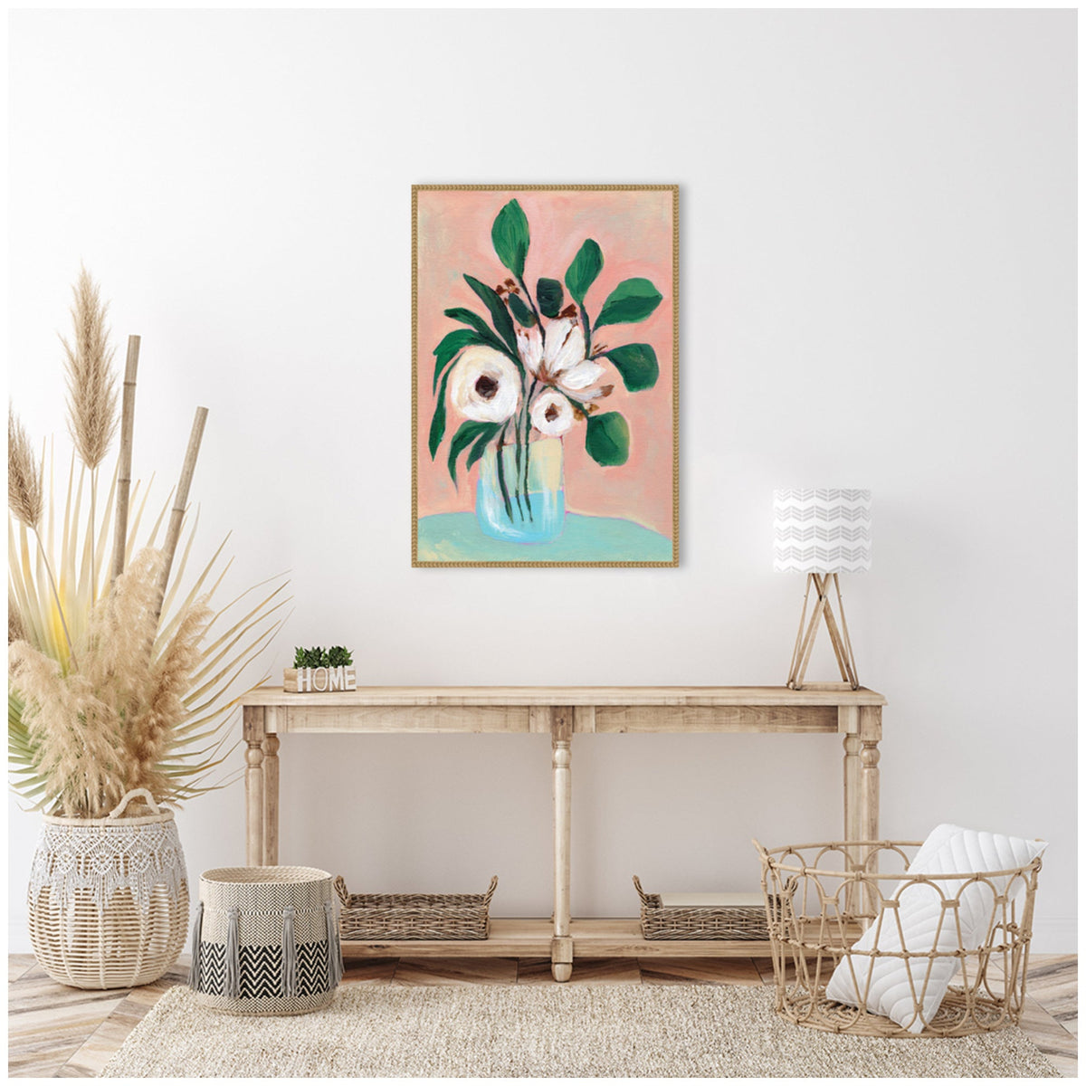 Framed floral painting with white flowers and dark green leaves on a pink and blue background, hanging above a light wood console table with wicker baskets and neutral decor.