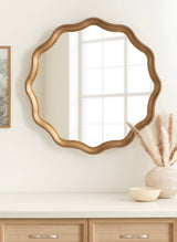 Round wall mirror with a scalloped, gold-toned frame reflecting a window, mounted above a light wood dresser with neutral decor including a vase and small bowl.