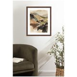 Abstract watercolor painting of muted earth-tone mountains with layered beige, brown, green, and black shapes in a dark wood frame hanging above an olive-green armchair and a potted plant.