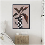 Abstract painting of large brown leaves in a tall black vase with white spots, framed and hanging above a rustic wooden bedside table with books and a cup.