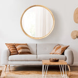 Round wood-framed wall mirror above a light gray sofa with three brown patterned cushions and a wood coffee table with white metal legs in a minimalist living room.