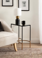 Rectangular side table with black metal frame and gold-toned lower legs, holding a black and brass lamp with white shade and a black mug beside a beige sofa.