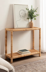 Rectangular rattan console table with woven texture, holding a line art portrait in a white frame and a beige vase with green leafy branches, on a wooden floor.